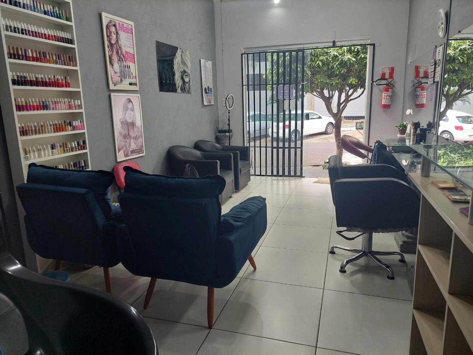 Recepção do salão com poltronas azuis, sofá de espera, vitrine de produtos e porta de vidro com vista para a rua